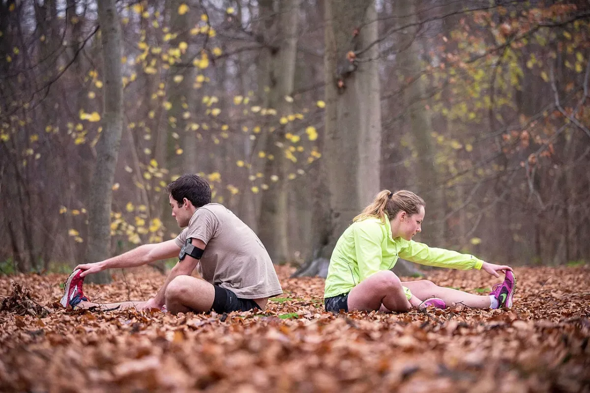 Two runners sitting on a leaf-covered forest floor, each reaching forward to hold the toes of an extended leg