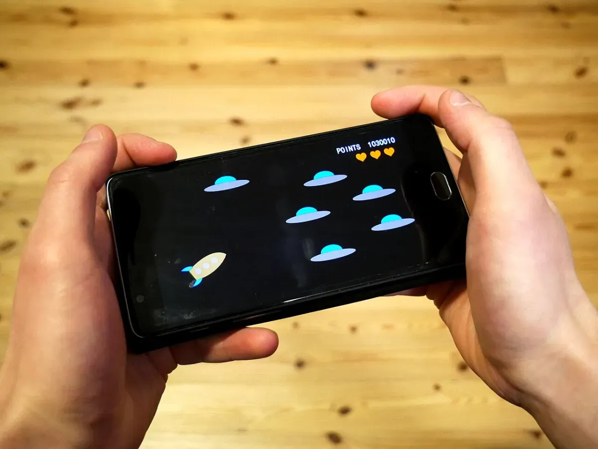 Two hands holding a smartphone sideways, playing a game at a wooden table