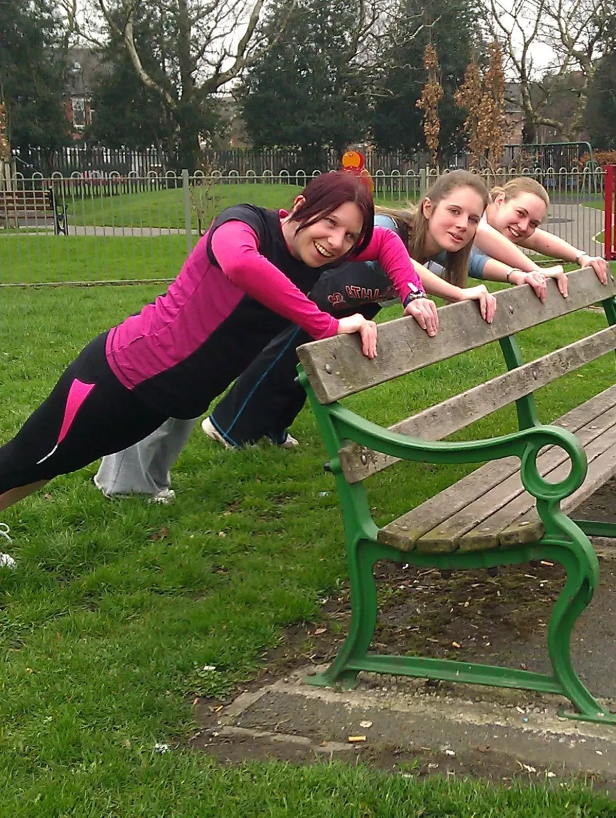 Three women doing incline push-ups with their hands on the backrest of a park bench, an easier push-up variation like a counter push-up