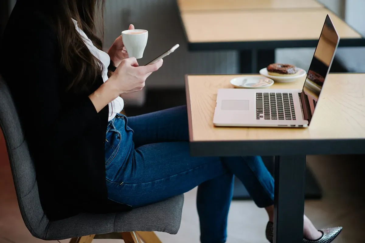 A woman at a cafe table checking her phone next to her laptop