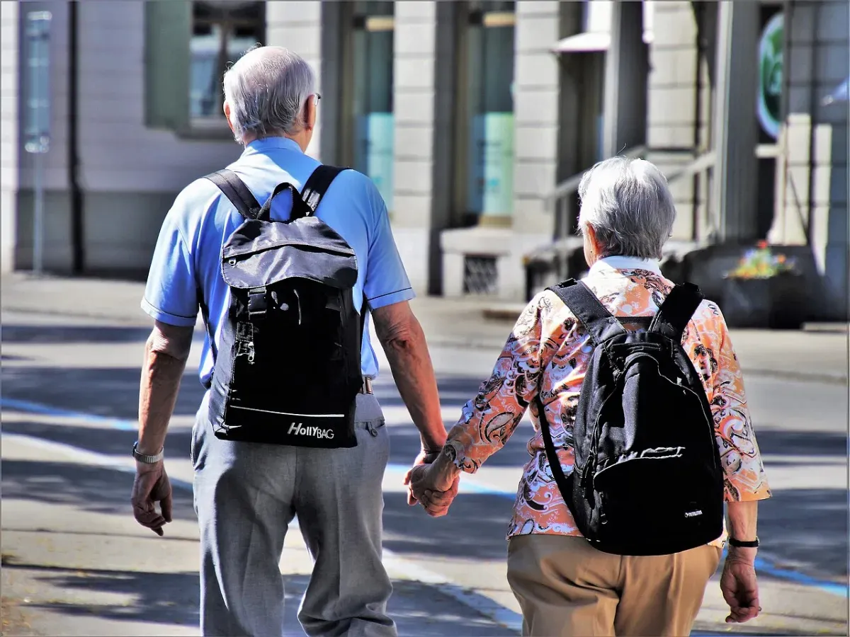 An older couple with backpacks walking hand in hand along a sunny sidewalk, seen from behind