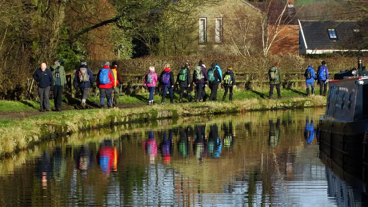 A group of walkers with backpacks on a canal towpath, reflected in the still water