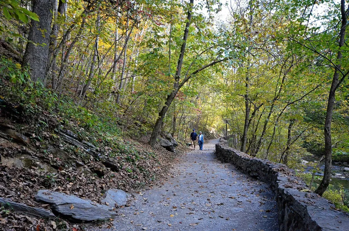 Two people walking along a gravel trail through autumn woods beside a stone wall