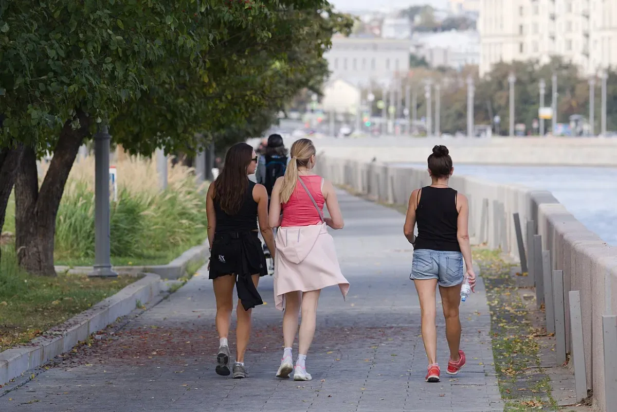 Three women in sneakers walking along a tree-lined riverside promenade, one carrying a water bottle