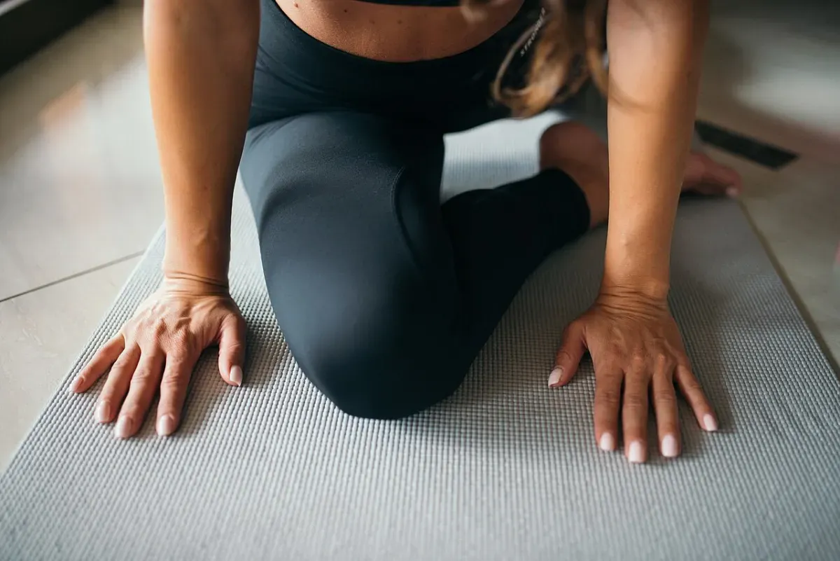 A woman kneeling on a grey yoga mat with one knee drawn forward and both palms on the floor, in a hip-opening position