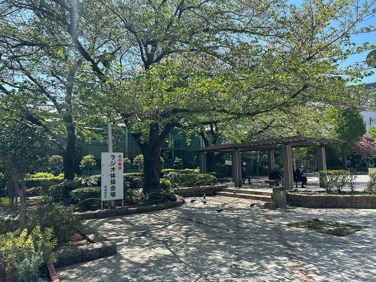 緑の多い公園の一角に立つ「年中無休 ラジオ体操会場」の看板